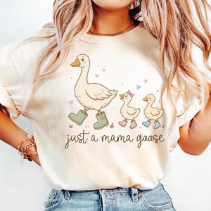 May include: Cream-colored t-shirt featuring a whimsical illustration of a goose and two goslings. The mama goose wears green boots, and the goslings wear pink and blue boots. The text "just a mama goose" is written below the illustration.