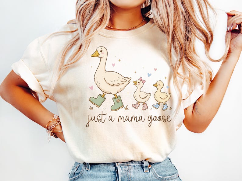 May include: Cream-colored t-shirt featuring a whimsical illustration of a goose and two goslings. The mama goose wears green boots, and the goslings wear pink and blue boots. The text "just a mama goose" is written below the illustration.