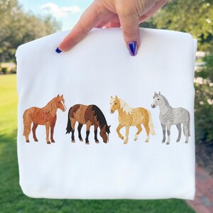 May include: White sweatshirt featuring four watercolor-style horses in shades of brown, tan, and gray. The horses are arranged horizontally across the front of the shirt, creating a nature-themed design.