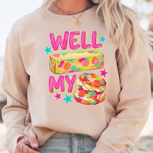 May include: A beige long-sleeve sweatshirt featuring a graphic print with the text WELL MY in bright pink. The design includes a yellow stick of butter with colourful paint-like splotches and a stack of two biscuits with colourful sprinkles and stars.
