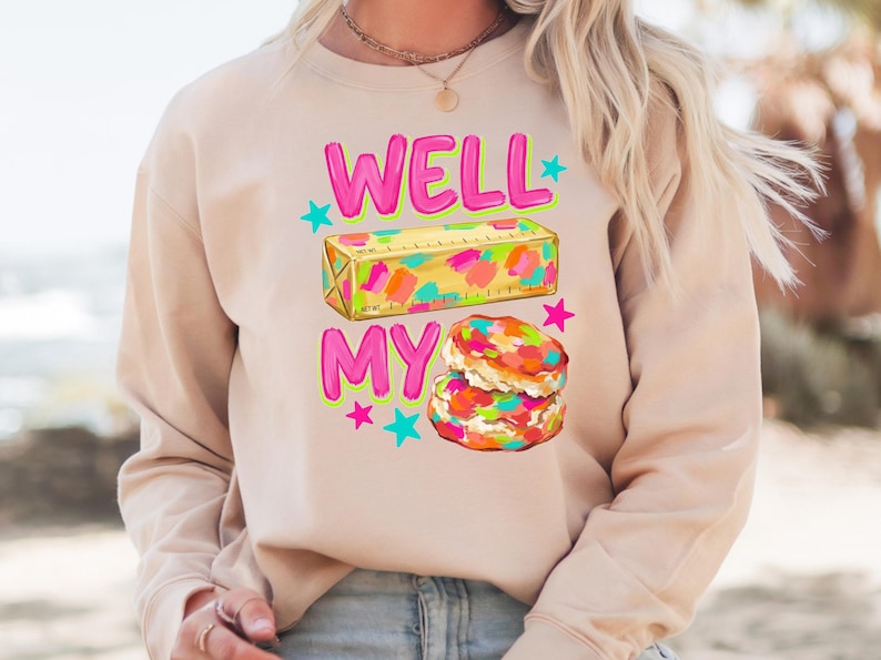 May include: A beige long-sleeve sweatshirt featuring a graphic print with the text WELL MY in bright pink. The design includes a yellow stick of butter with colourful paint-like splotches and a stack of two biscuits with colourful sprinkles and stars.