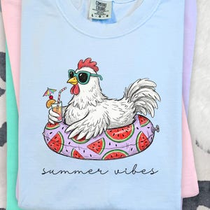 May include: Light blue t-shirt with a graphic of a white chicken wearing sunglasses and floating on a watermelon-themed inflatable ring. The chicken holds a cocktail. The words "summer vibes" are written below the graphic.
