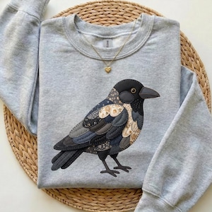 Può includere: Una felpa grigio chiaro con un'applicazione dettagliata di un corvo. Il design del corvo incorpora varie tonalità di grigio, nero e beige. La felpa ha un girocollo e maniche lunghe. È visibile anche una collana d'oro con un ciondolo a forma di cuore.