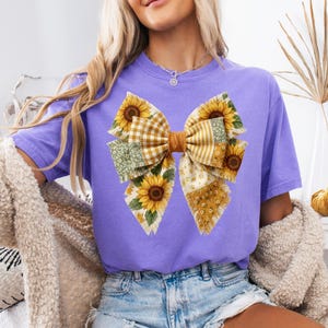 May include: A lilac-colored t-shirt featuring a large bow design with sunflowers and patchwork patterns. The bow has a brown center and is adorned with yellow sunflowers, green floral, and plaid fabrics. The shirt is paired with denim shorts.