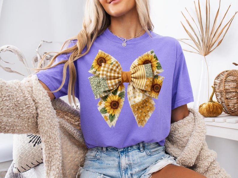 May include: A lilac-colored t-shirt featuring a large bow design with sunflowers and patchwork patterns. The bow has a brown center and is adorned with yellow sunflowers, green floral, and plaid fabrics. The shirt is paired with denim shorts.