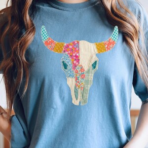 May include: A light blue t-shirt featuring a colorful patchwork bull skull design. The skull is adorned with various floral and patterned fabrics in shades of pink, yellow, blue, and green. The shirt is a comfortable, casual style.
