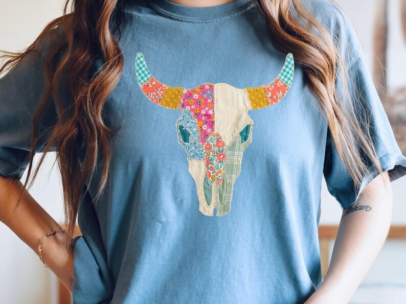 May include: A light blue t-shirt featuring a colorful patchwork bull skull design. The skull is adorned with various floral and patterned fabrics in shades of pink, yellow, blue, and green. The shirt is a comfortable, casual style.