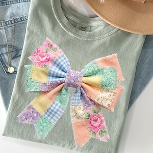 Pu&ograve; includere: Una maglietta verde chiaro con un grande fiocco patchwork colorato. Il fiocco presenta toppe floreali, a quadretti e in tinta unita in colori pastello. Sono visibili anche jeans azzurri e un cappello beige.