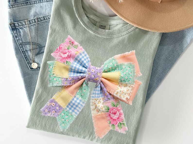 Pu&ograve; includere: Una maglietta verde chiaro con un grande fiocco patchwork colorato. Il fiocco presenta toppe floreali, a quadretti e in tinta unita in colori pastello. Sono visibili anche jeans azzurri e un cappello beige.