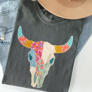May include: A gray t-shirt featuring a colorful patchwork bull skull design. The shirt is paired with light-wash denim jeans, a tan hat, and a silver necklace. The bull skull has floral and patterned accents.