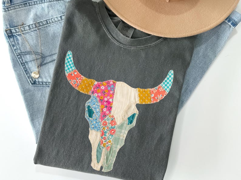 May include: A gray t-shirt featuring a colorful patchwork bull skull design. The shirt is paired with light-wash denim jeans, a tan hat, and a silver necklace. The bull skull has floral and patterned accents.