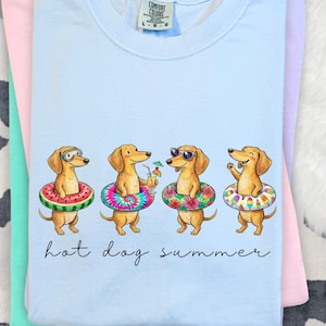 May include: A light blue t-shirt with a graphic of four dachshunds wearing inflatable rings and accessories. The phrase "hot dog summer" is written below. The shirt is folded with other pastel coloured shirts. The t-shirt is a Comfort Colors brand.