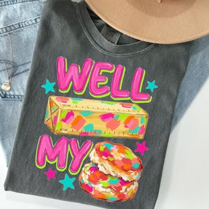 May include: A charcoal grey t-shirt featuring the text WELL MY in bright pink with a yellow stick of butter graphic decorated with colourful paint splatters and two stacked biscuits with similar colourful splatters, accented by small pink and teal stars.