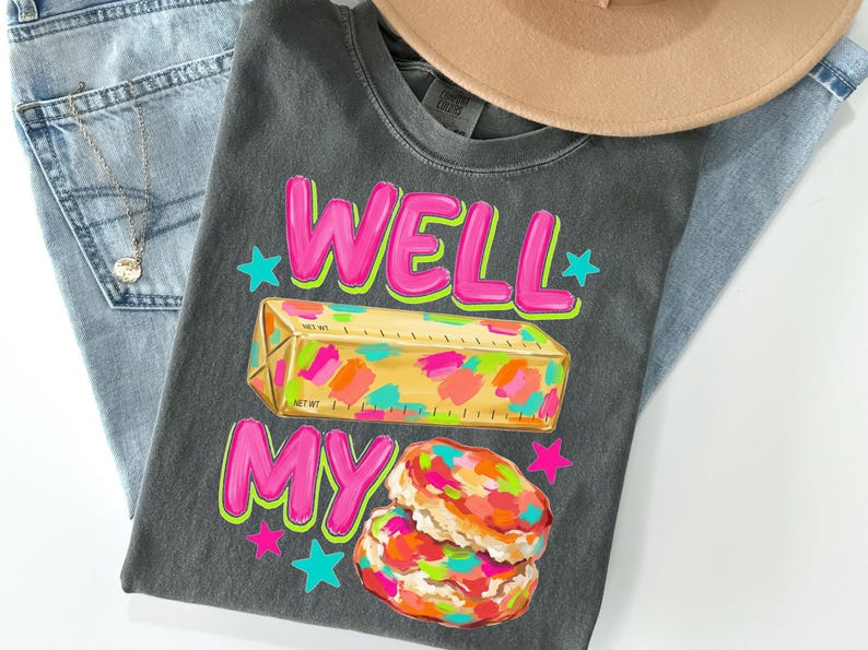 May include: A charcoal grey t-shirt featuring the text WELL MY in bright pink with a yellow stick of butter graphic decorated with colourful paint splatters and two stacked biscuits with similar colourful splatters, accented by small pink and teal stars.