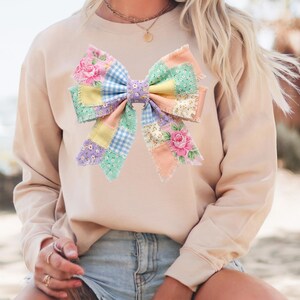 Pu&ograve; includere: Una felpa beige con un'applicazione a fiocco grande e colorata. Il fiocco &egrave; realizzato in tessuto patchwork nei colori pastello, tra cui fiori rosa, quadretti blu e quadrati gialli e verdi a tinta unita. Il fiocco &egrave; il punto focale del top casual.