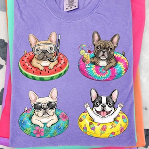 May include: A purple t-shirt featuring four French bulldogs in inflatable rings. One dog wears goggles and a snorkel, another holds a cocktail, a third wears sunglasses, and the last one is smiling. The rings have watermelon, tie-dye, floral, and popsicle designs.