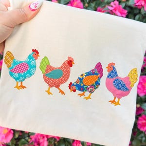 May include: A cream-colored tote bag featuring four colorful chicken appliqués. Each chicken has a unique pattern, including floral, polka dot, and plaid designs. The chickens are in shades of blue, red, pink, and yellow, with orange feet and red combs.
