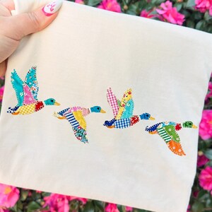 May include: Cream-colored fabric featuring four embroidered ducks in flight. Each duck has a unique patchwork design with vibrant colors like blue, yellow, pink, and green. The ducks are flying across the fabric, creating a dynamic and whimsical scene.