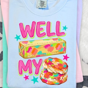 May include: A light blue t-shirt featuring a graphic of a stick of butter and two biscuits with colourful paint-like splatters. The text reads WELL MY in bright pink letters with a neon green outline, accented by small pink and teal stars.