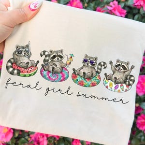 May include: A cream-colored tote bag featuring four raccoons relaxing on inflatable rings. Each raccoon is depicted with sunglasses or a snorkel, and the text "feral girl summer" is written in a script font. The background is a vibrant display of pink flowers.