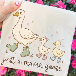 May include: A cream-colored tote bag featuring a cartoon illustration of a goose and two goslings, all wearing boots. The text "just a mama goose" is written in a whimsical font. Pink flowers and hearts are scattered around the design.