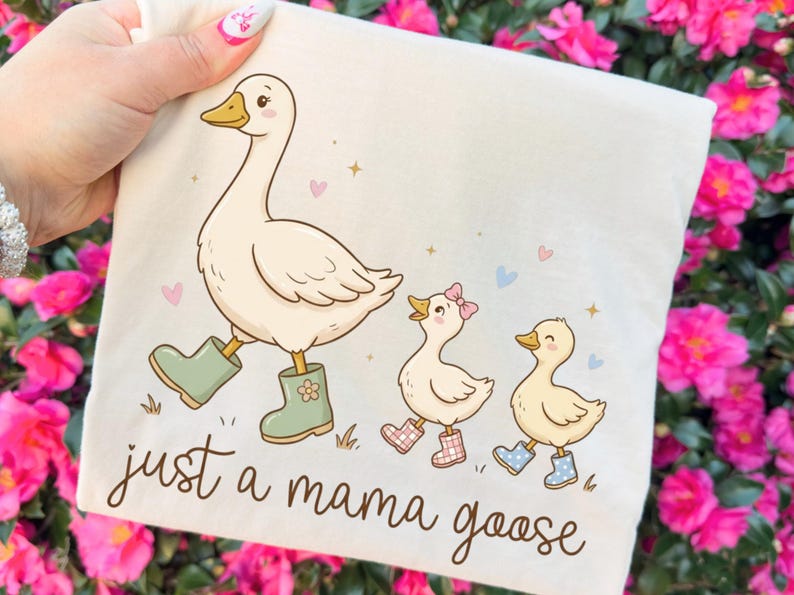 May include: A cream-colored tote bag featuring a cartoon illustration of a goose and two goslings, all wearing boots. The text "just a mama goose" is written in a whimsical font. Pink flowers and hearts are scattered around the design.