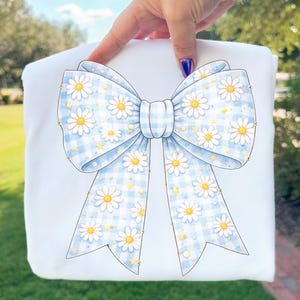 May include: A white item featuring a large bow design. The bow has a light blue gingham pattern with white daisies and yellow centers. The bow is centered on the item, with the background out of focus.