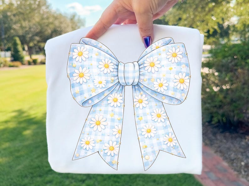 May include: A white item featuring a large bow design. The bow has a light blue gingham pattern with white daisies and yellow centers. The bow is centered on the item, with the background out of focus.