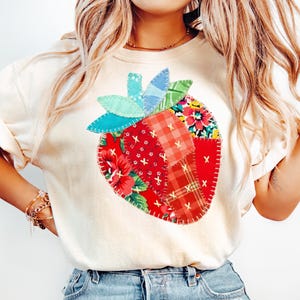 May include: Cream-colored t-shirt featuring a large, patchwork strawberry design. The strawberry is composed of various fabric patterns, including red floral, plaid, and solid red pieces. The stem and leaves are made of blue and green fabric.