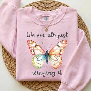 May include: A light pink sweatshirt with the text "We are all just winging it" and a colorful butterfly design. The butterfly has orange, red, yellow, green, and blue wings. A gold necklace with a heart pendant is also visible.