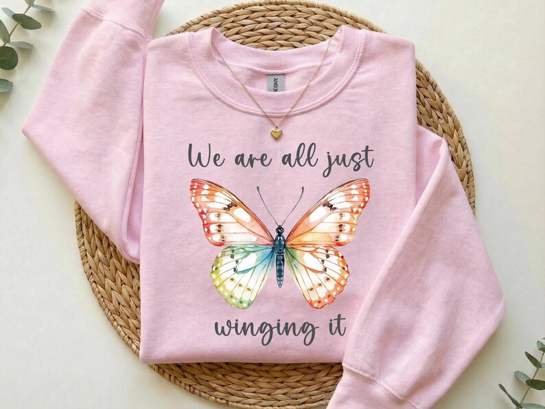 May include: A light pink sweatshirt with the text "We are all just winging it" and a colorful butterfly design. The butterfly has orange, red, yellow, green, and blue wings. A gold necklace with a heart pendant is also visible.