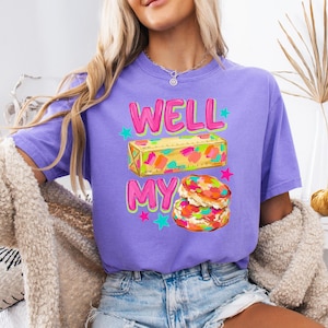 May include: A purple short-sleeve t-shirt featuring a graphic print with the text WELL MY. The design includes a yellow stick of butter with colourful paint splatters and a stack of two biscuits with colourful sprinkles, accented by small teal and pink stars.