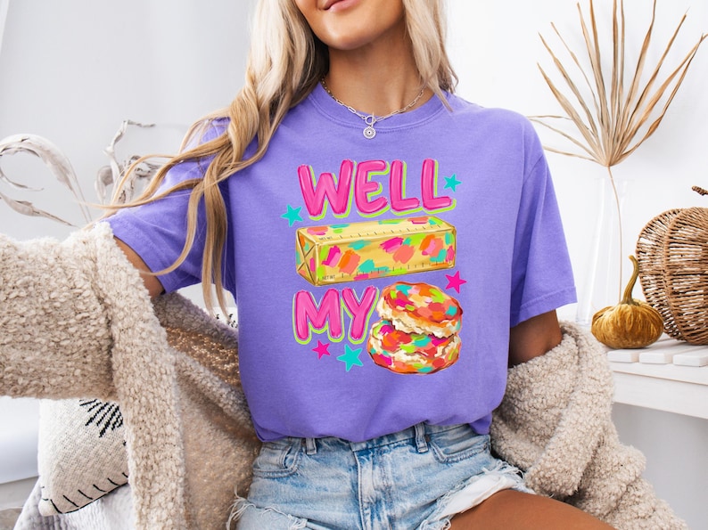 May include: A purple short-sleeve t-shirt featuring a graphic print with the text WELL MY. The design includes a yellow stick of butter with colourful paint splatters and a stack of two biscuits with colourful sprinkles, accented by small teal and pink stars.