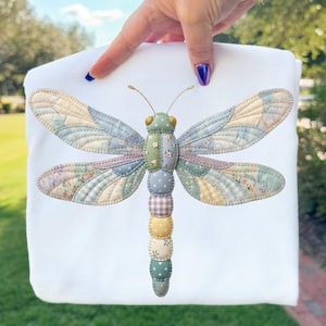 May include: A white fabric item featuring a large, colorful dragonfly applique. The dragonfly has patchwork wings in pastel shades of blue, yellow, and green, with a patterned body. The item is held up by a hand with blue nail polish.