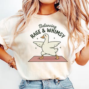 Pode incluir: Camiseta creme com a frase "Balancing Rage & Whimsy" e um desenho de um ganso em posição de ioga. O ganso branco tem as asas abertas, sobre um tapete de ioga rosa. A camiseta tem mangas curtas.