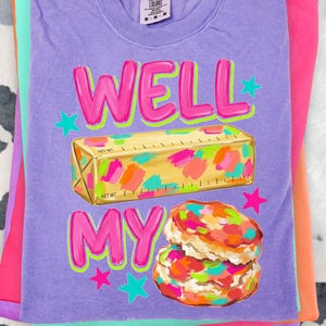 May include: A purple short-sleeve t-shirt featuring a graphic print of a stick of butter with colourful paint-like splatters and a stack of two biscuits with similar colourful splatters. The text WELL MY reads in bright pink with green outlines.