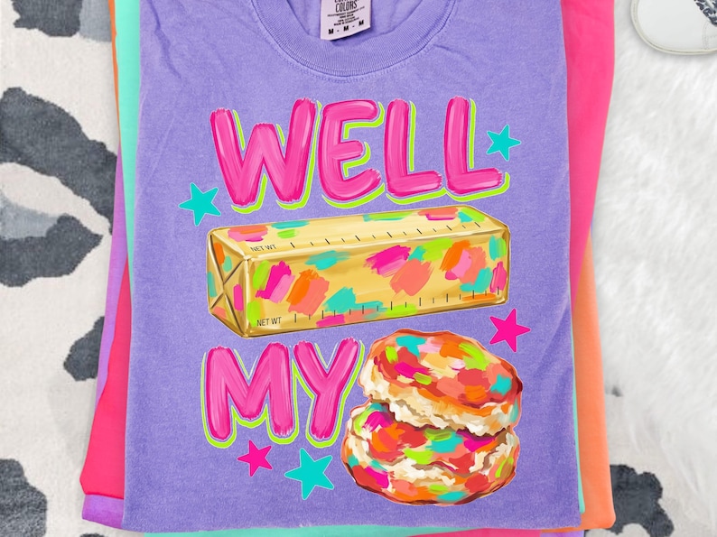 May include: A purple short-sleeve t-shirt featuring a graphic print of a stick of butter with colourful paint-like splatters and a stack of two biscuits with similar colourful splatters. The text WELL MY reads in bright pink with green outlines.
