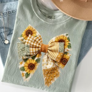 May include: A light green t-shirt featuring a large bow design with sunflowers and various patterned fabrics. The bow includes yellow gingham, floral, and striped patterns. A pair of blue jeans and a tan hat are also in the image.