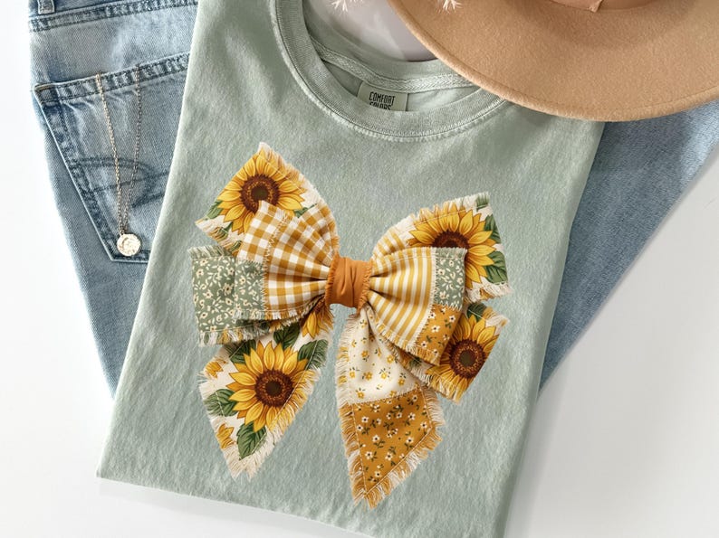 May include: A light green t-shirt featuring a large bow design with sunflowers and various patterned fabrics. The bow includes yellow gingham, floral, and striped patterns. A pair of blue jeans and a tan hat are also in the image.