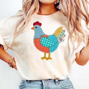 Könnte beinhalten: Cremefarbenes T-Shirt mit einem farbenfrohen Patchwork-Hühnerdesign. Das Huhn hat einen roten Kamm, einen orangefarbenen Schnabel und besteht aus verschiedenen gemusterten Stoffen in Blau, Orange und Gelb. Das Shirt ist ein lässiger Stil.