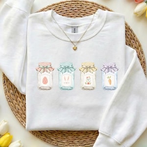 May include: White sweatshirt featuring four decorative jars with Easter-themed illustrations. Each jar has a different design: an egg, bunny ears, flowers, and a carrot. The jars are adorned with colorful bows and are set against a neutral background.