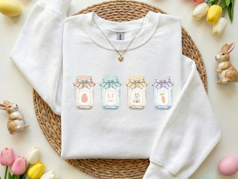 May include: White sweatshirt featuring four decorative jars with Easter-themed illustrations. Each jar has a different design: an egg, bunny ears, flowers, and a carrot. The jars are adorned with colorful bows and are set against a neutral background.