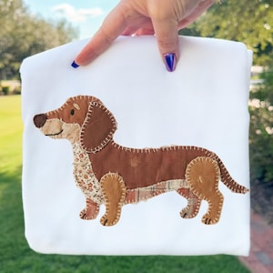 May include: A white fabric item with a brown dachshund applique. The dog has a patchwork design with floral and plaid patterns, and is stitched with white thread. The item is held up by a hand with blue nail polish.