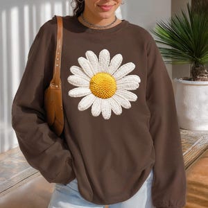 May include: A brown sweatshirt featuring a large daisy design. The daisy has white petals and a yellow center. The sweatshirt is a solid color and has long sleeves. The overall style is casual and comfortable.