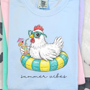 May include: Light blue t-shirt featuring a cartoon chicken wearing sunglasses and floating on a yellow and teal inner tube, holding a tropical drink. The words "summer vibes" are written in black script below the image. The shirt is folded with other pastel colored shirts.