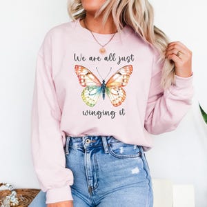 May include: A light pink sweatshirt with a colorful butterfly graphic. The butterfly has orange, yellow, blue, and green wings. The text "We are all just winging it" is above and below the butterfly.
