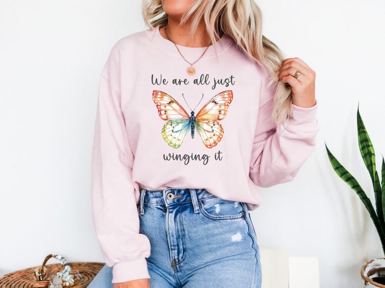 May include: A light pink sweatshirt with a colorful butterfly graphic. The butterfly has orange, yellow, blue, and green wings. The text "We are all just winging it" is above and below the butterfly.