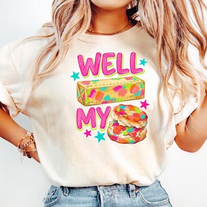 May include: A cream-coloured t-shirt featuring a graphic print with the text WELL MY in bright pink letters. The design includes a yellow stick of butter with colourful paint-like splotches and a stack of two biscuits with similar colourful accents.