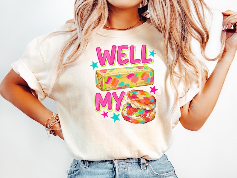 May include: A cream-coloured t-shirt featuring a graphic print with the text WELL MY in bright pink letters. The design includes a yellow stick of butter with colourful paint-like splotches and a stack of two biscuits with similar colourful accents.