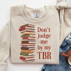 May include: A beige sweatshirt with the text "Don't judge me by my TBR" in red. A stack of books in various colors is printed on the left side. The sweatshirt is paired with blue jeans and a brown mug.